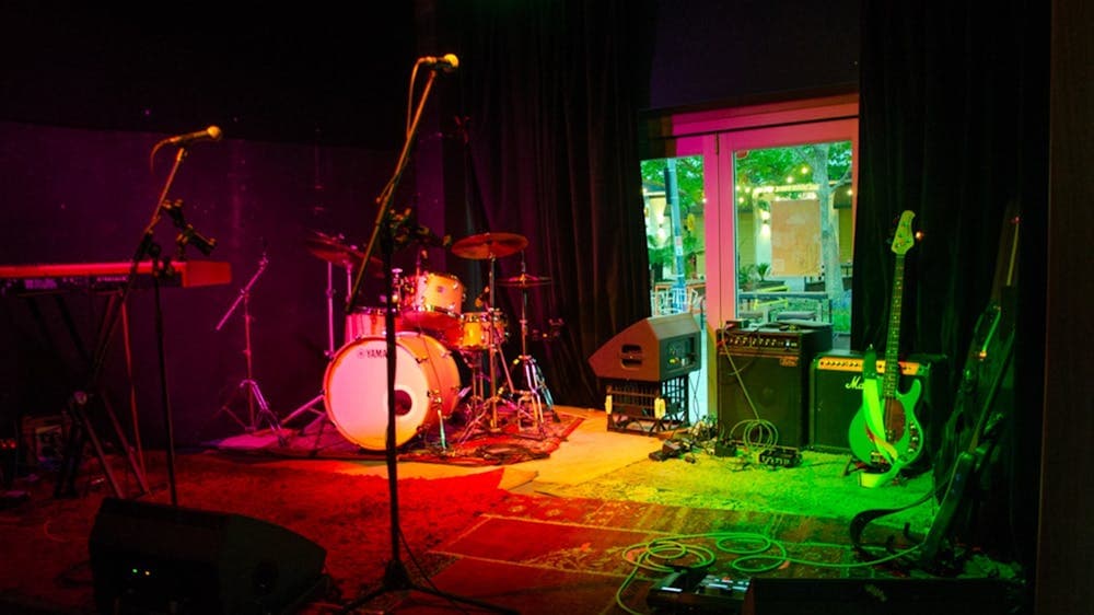 Image of a drumset, microphone, a couple of amps on  a stage with red and green lighting.
