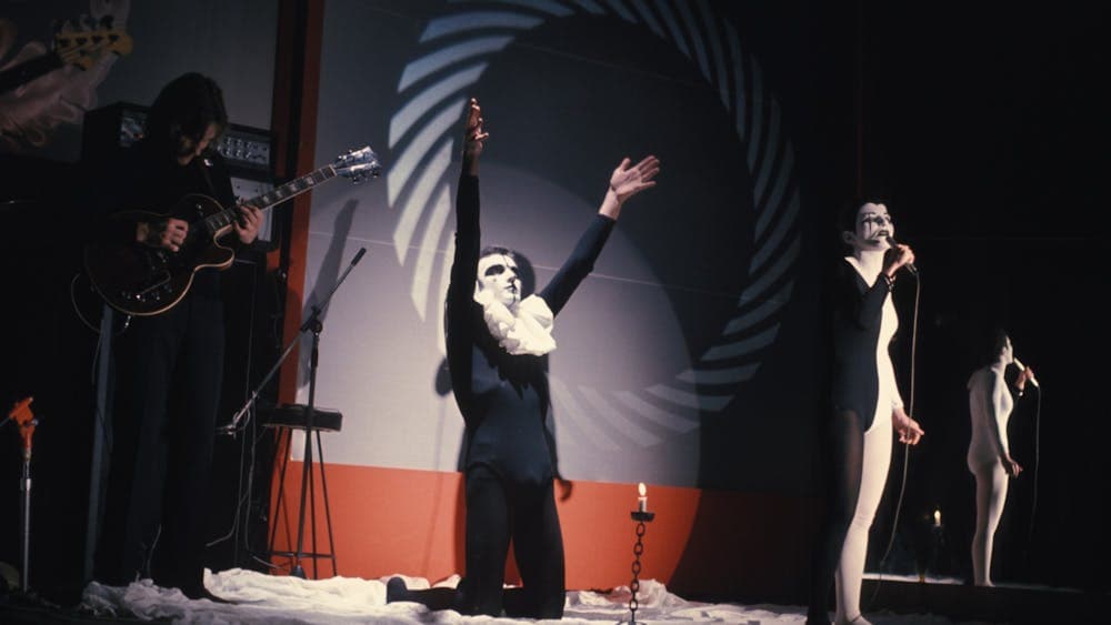 Band on stage wearing black and white costumes with main singer on his knees