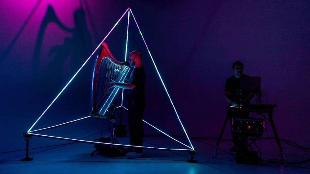 A harpist performs inside a glowing triangular light structure, operated by a man in the shadows