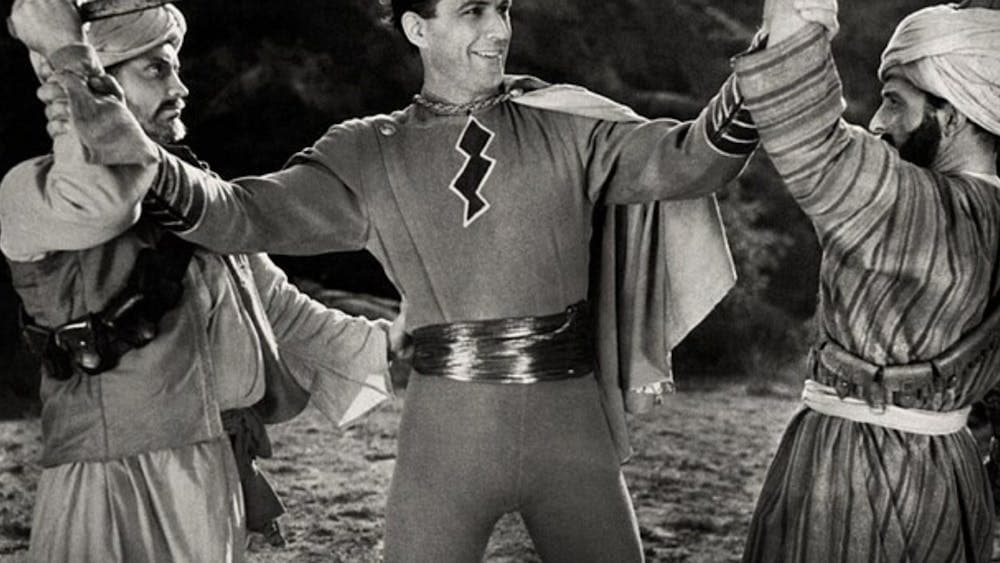 black and white image of man dressed as a superhero smiling, with two people on either side