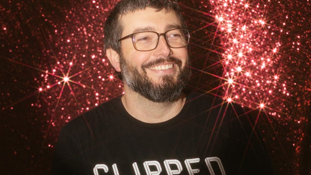 Male with glasses looks past the camera smiling with a sparkly background