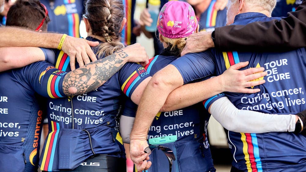 Signature Tour cyclists in kit, in a huddle with arms around each other, taken from behind them